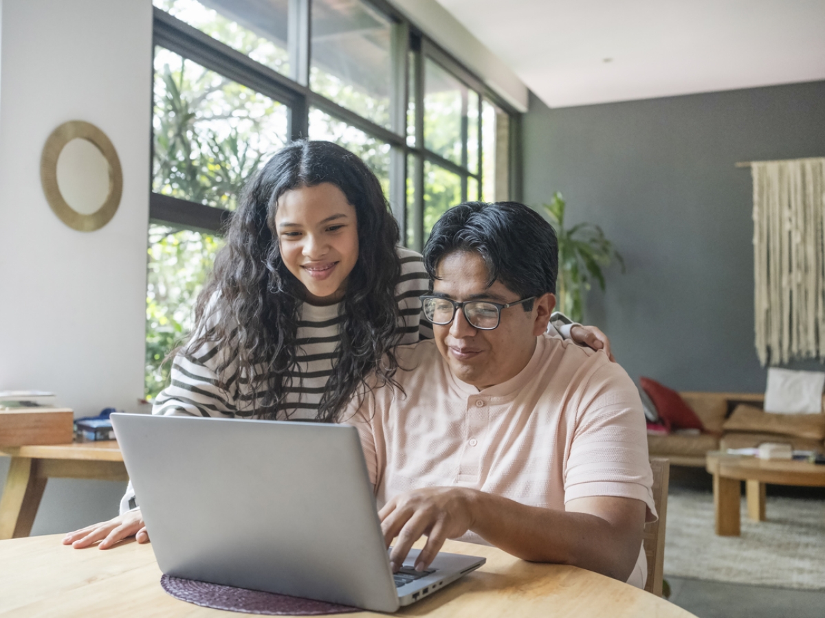 Young woman standing over adult man's shoulder, both looking at a laptop screen.